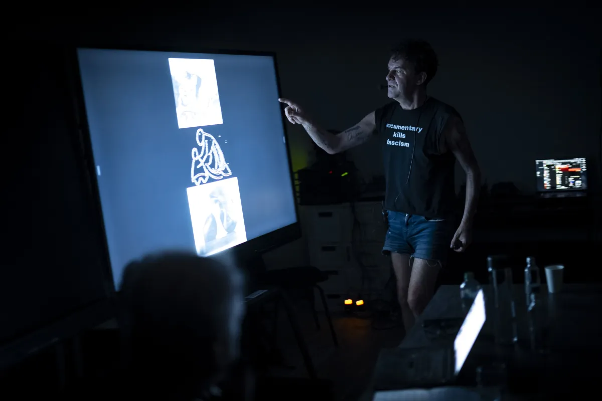A skinny middle-aged white man in short shorts and a t-shirt that reads "Documentary Kills Fascism" stands in a darkened room pointing at a screen with a few film images