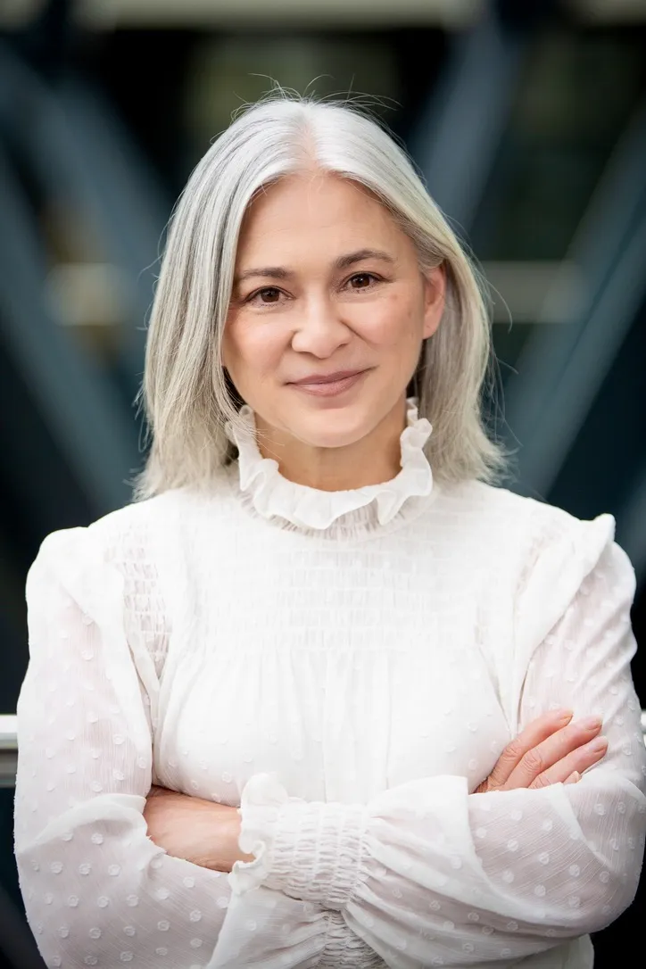 Smiling woman with white hair in a frilly white blouse crosses her arms and looks into the camera in front of a dark blue out-of-focus background