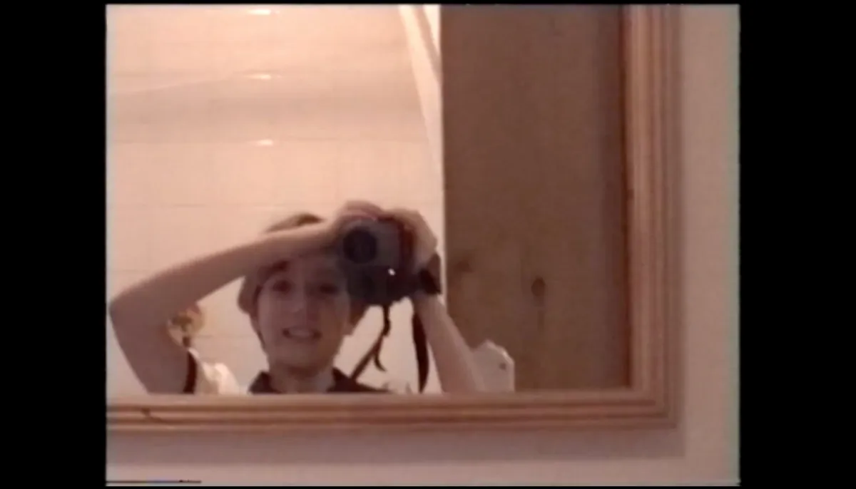 Pink tinted image of a smiling little girl with short hair filming herself in a mirror
