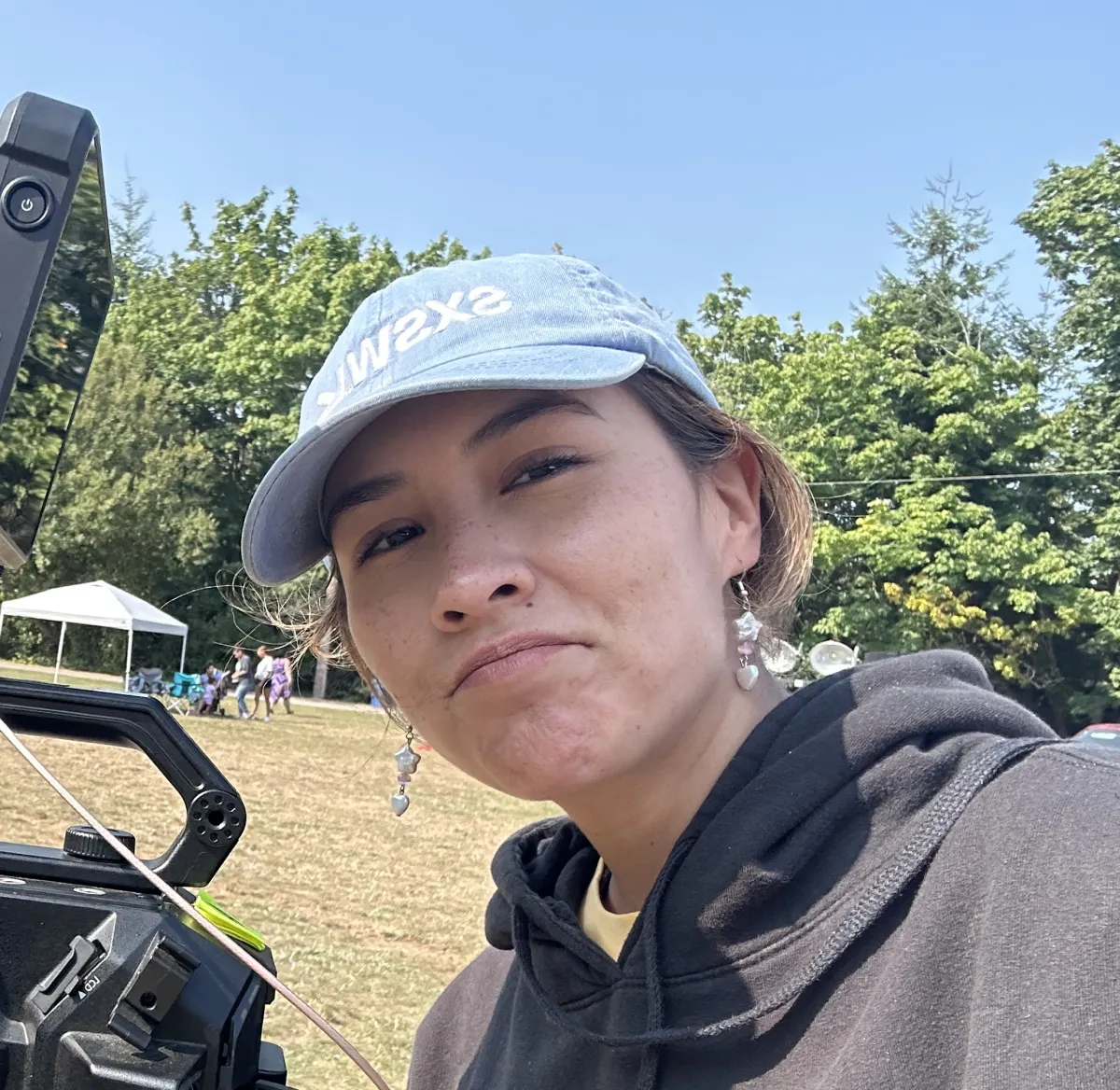 Young Indigenous woman with her hair pulled back under a light blue ballcap wears a dark hoodie as she takes a selfie outside, a film camera to her right