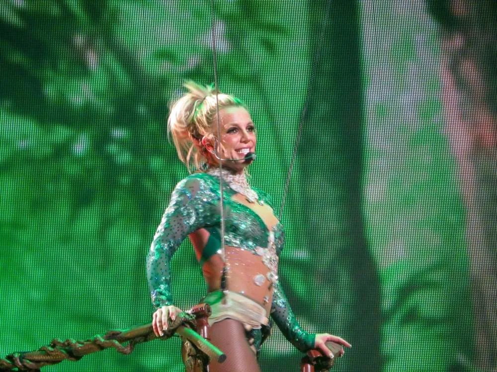 Pop star Britney Spears, subject the recnt documentary 'Framing Britney Spears," is pictured performing on stage, wearing an emerald green sequin outfit, singing into a wireless headset, clutching a brown railing, against a mesh, emerald green patterned backdrop.