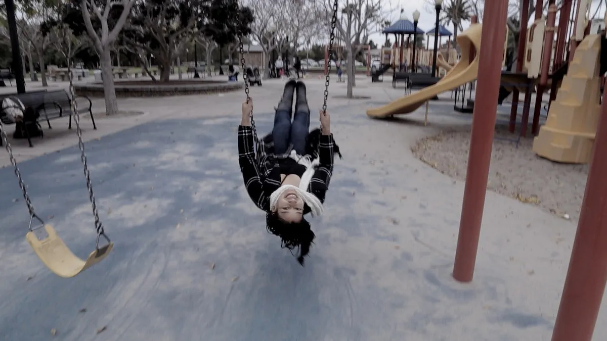 A woman from the film swings on  a swing in the middle of a playground; her head is upside-down as she faces the camera.