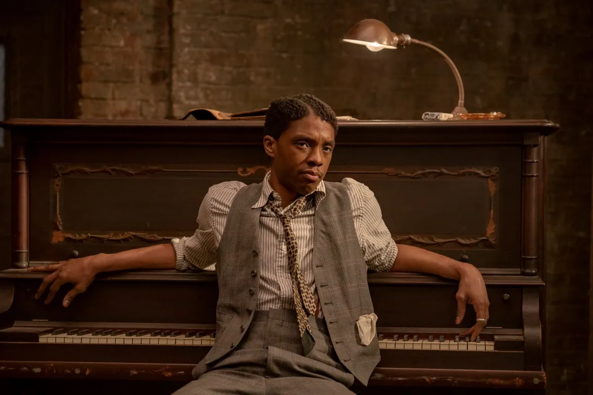 Actor Chadwick Boseman, a Black man wearing a striped shirt, patterned tie, grey vest and pants, sits at a piano, his back to the keyboard