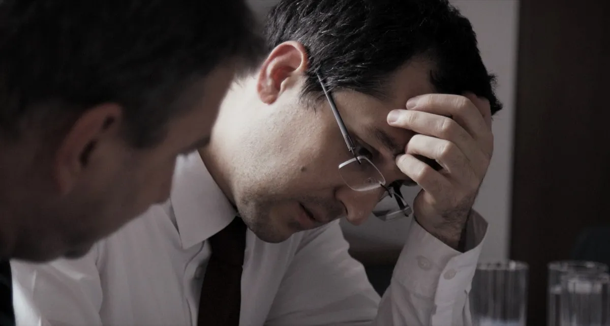 The image shows a close-up profile of a government official, sitting at a desk with his colleagues, examining documents. He has dark hair and glasses and is wearing a white shirt and a dark tie.