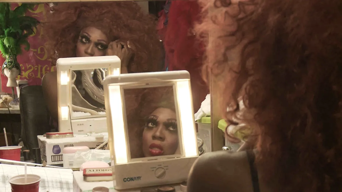 BeBe Zahara Benet is a Black drag queen preparing for a show in her dressing room in Minneapolis. She is wearing a very large brown wig and false eyelashes, while looking into a mirror. From Emily Branham’s ‘Being BeBe.’ Courtesy of the filmmaker.