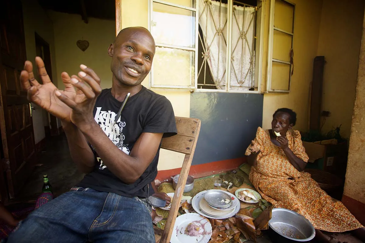 David Kato, a Black man with a bald head sits in front of a yellow house. His mother, Lydia Mulumba Nalongo, an older Black woman sits behind him eating. A still from 'Call Me Kutchu,' courtesy of the filmmakers.
