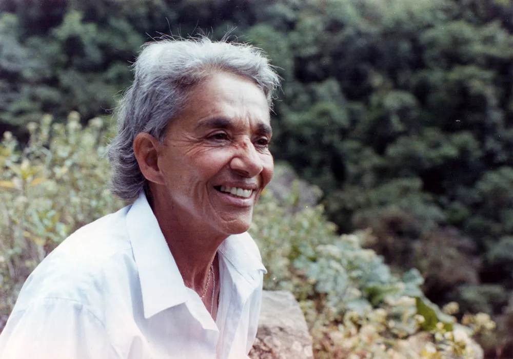 Chavela Vargas is a queer Latin American singer with grey hair. She is wearing a white shirt and is smiling. She is surrounded by trees and shrubs. Image from Catherine Gund and Daresha Kyi’s ‘Chavela.’ Courtesy of Alicia Elena Pérez Duarte, Aubin Pictures.