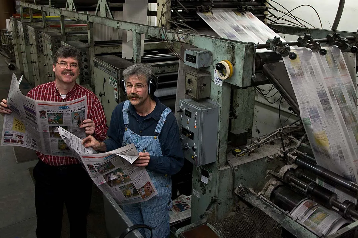 Brothers John and Art Cullen are two white men standing in a printing press, reviewing a hot-off-the-presses copy of 'The Storm Lake Times.' Image from Beth Levison and Jerry Risius’ ‘Storm Lake.’ Courtesy of 'Independent Lens'/PBS. 