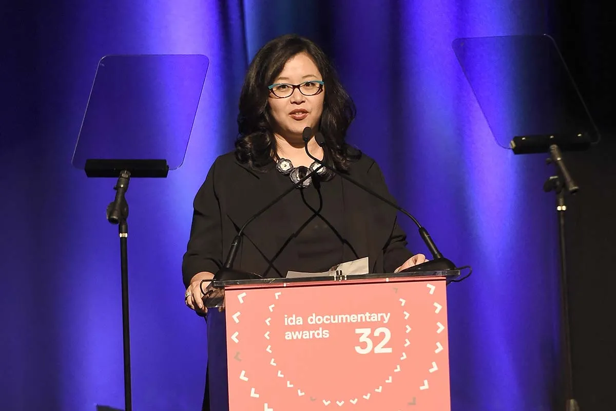 MacArthur Foundation's Kathy Im is an Asian American woman with shoulder-length black hair and glasses, wearing a black dress and standing at the podium at the IDA Documentary Awards.