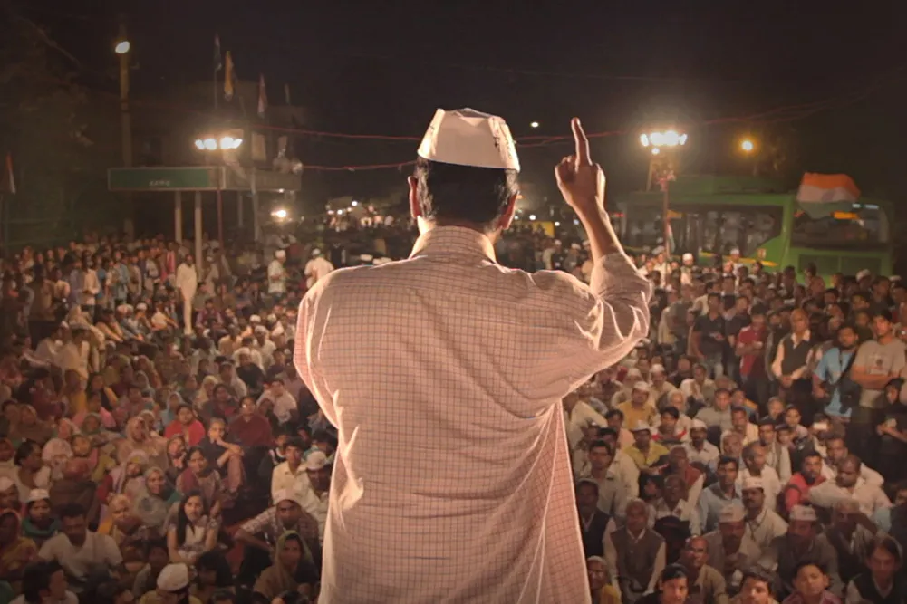 A man with brown skin in a white checkered shirt and white hat standing in front of a crowd. Still from 'An Insignificant Man' by Khushboo Ranka & Vinay Shukla. Courtesy of IFFLA. 