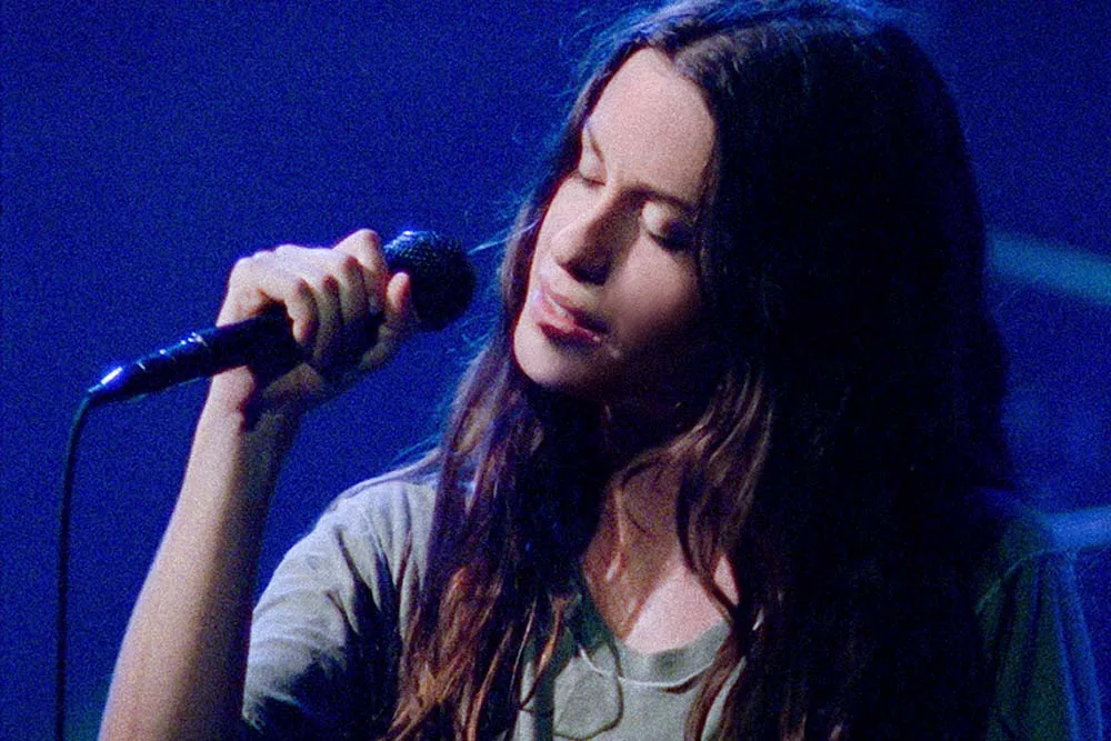 Singer Alanis Morissette, a white woman with long black hair, is performing on stage against a blue lit background; she is holding a microphone, and her eyes are closed, From Alison Kaynman’s ‘Jagged.’ Courtesy of TIFF.