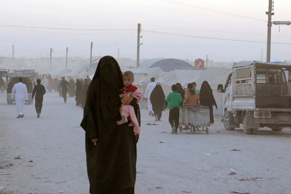  A woman in a burqa, her face covered, standing with a child on her lap. There are people walking behind her. Image from Hogir Hirori’s 'Sabaya.' Courtesy of Susan Norget Film Promotion.