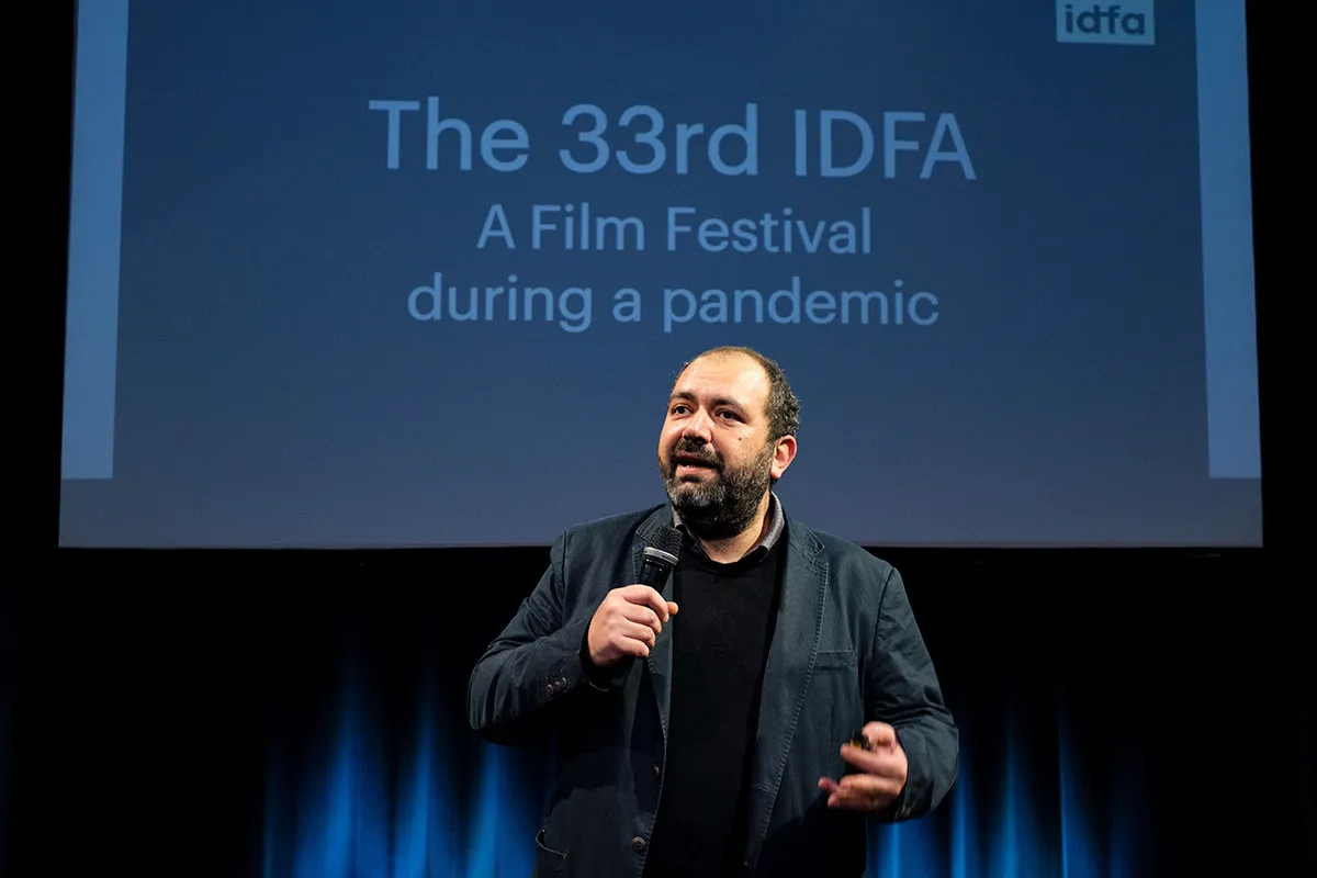 IDFA Artistic Director and filmmaker Orwa Nyrabia is a Syrian man with a beard and short hair. He is wearing a black jacket and a sweater while addressing the in-person and virtual audience at IDFA 2020. There is a screen behind him. Photo: Coen Dijkstra. Courtesy of IDFA