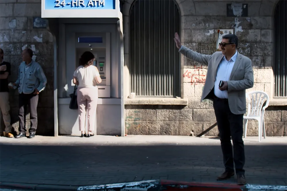 Musa Hadid, the mayor of Ramallah is a middle-aged man with a moustache. He is waving to someone and is wearing sunglasses, and is wearing a grey jacket with a shirt and trousers. There are passersby and an ATM machine behind him. Image from David Osit’s 'Mayor.' Courtesy of David Osit and POV.