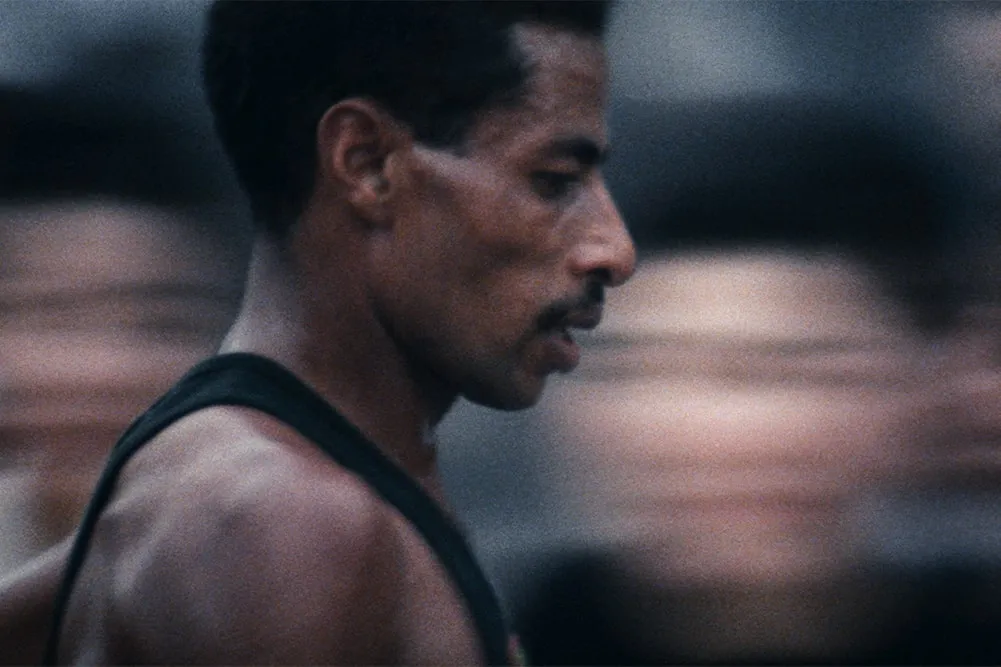 Abebe Bikila is a Black Ethiopian marathoner seen here at the 1964 Summer Olympics at Tokyo. He has black hair, a moustache, and is wearing a black vest. Image from Kon Ichikawa’s ‘Tokyo Olympiad.’ Courtesy of The Criterion Collection.