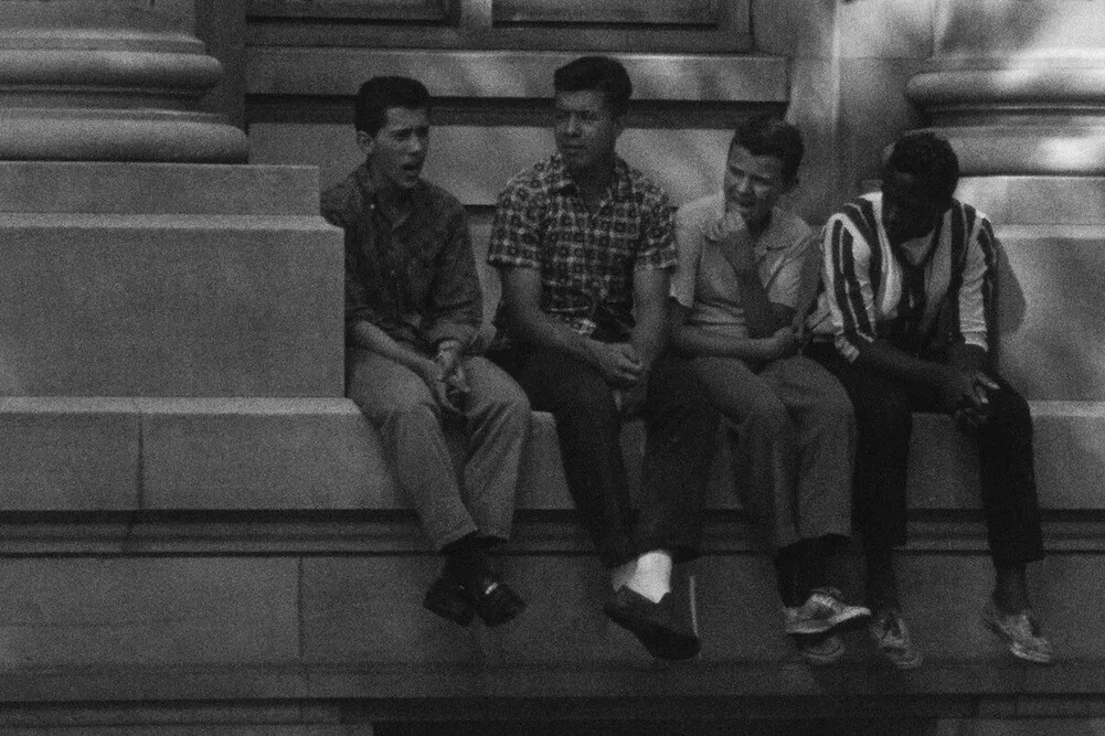 A BW photo of four young boys—one Black, three white—sitting on a building ledge