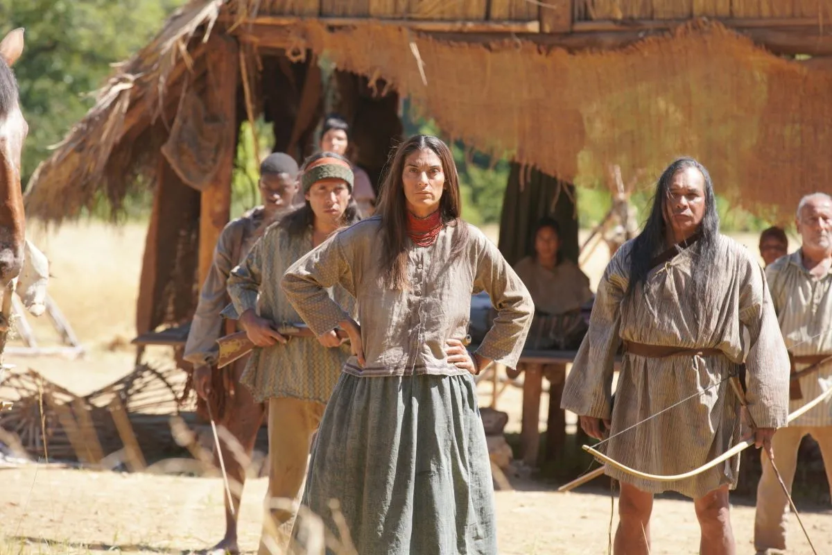 A group of 19th century Seminole tribespeople, wearing long-sleeved tunics and armed with bows & arrows, guard their dwelling.