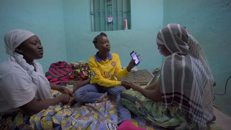 Three African girls sit in a room with a turquoise background as one shows her phone. From Ousmane Samassékou’s 'The Last Shelter.' Photo courtesy of STEPS. 