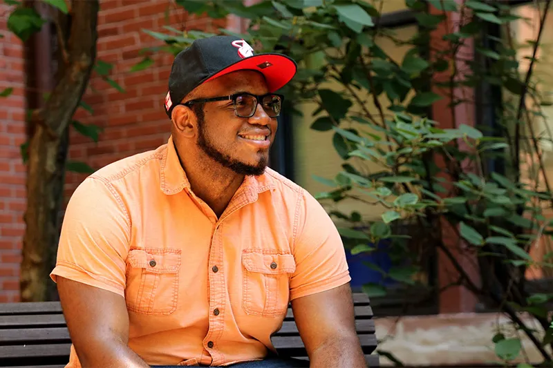   Headshot of a smiling Black adult man sitting on a bench in a black and red hat, glasses, and an orange button-up shirt.