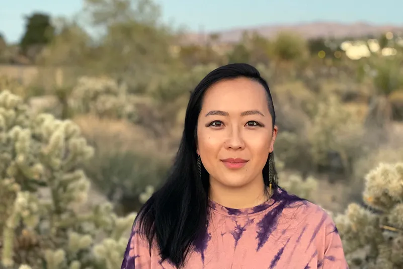 Asian-American adult is standing in front of desert landscape, wearing pink tie-dye tee, black hair passed the shoulders with left side shaved.