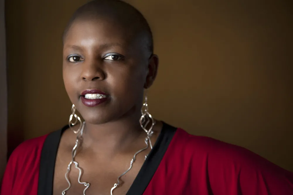 Yoruba Richen is an African American woman. She has a shaved head and is sporting large abstract dangled metal earrings that reaches her chest. She is wearing a red V-neck top with contrasting black neck band.