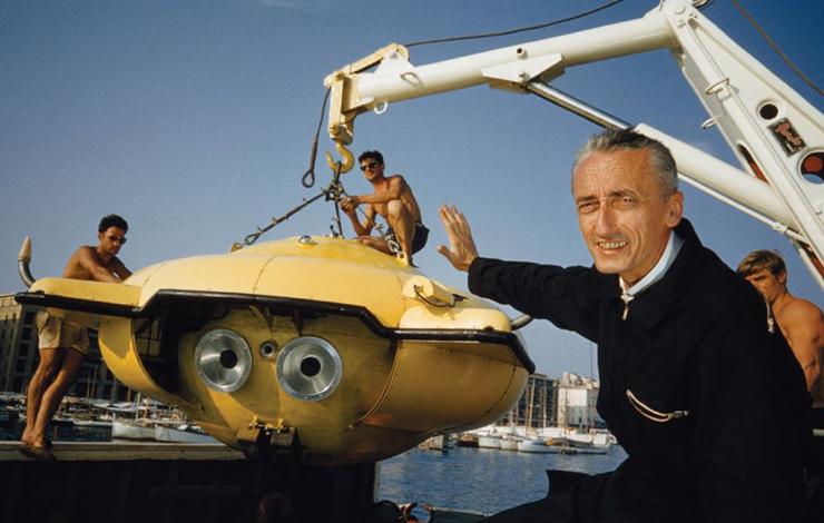 An image of documentary filmmaker Jacques-Yves Cousteau, an older white man with short gray hair, standing in front of a yellow submarine.