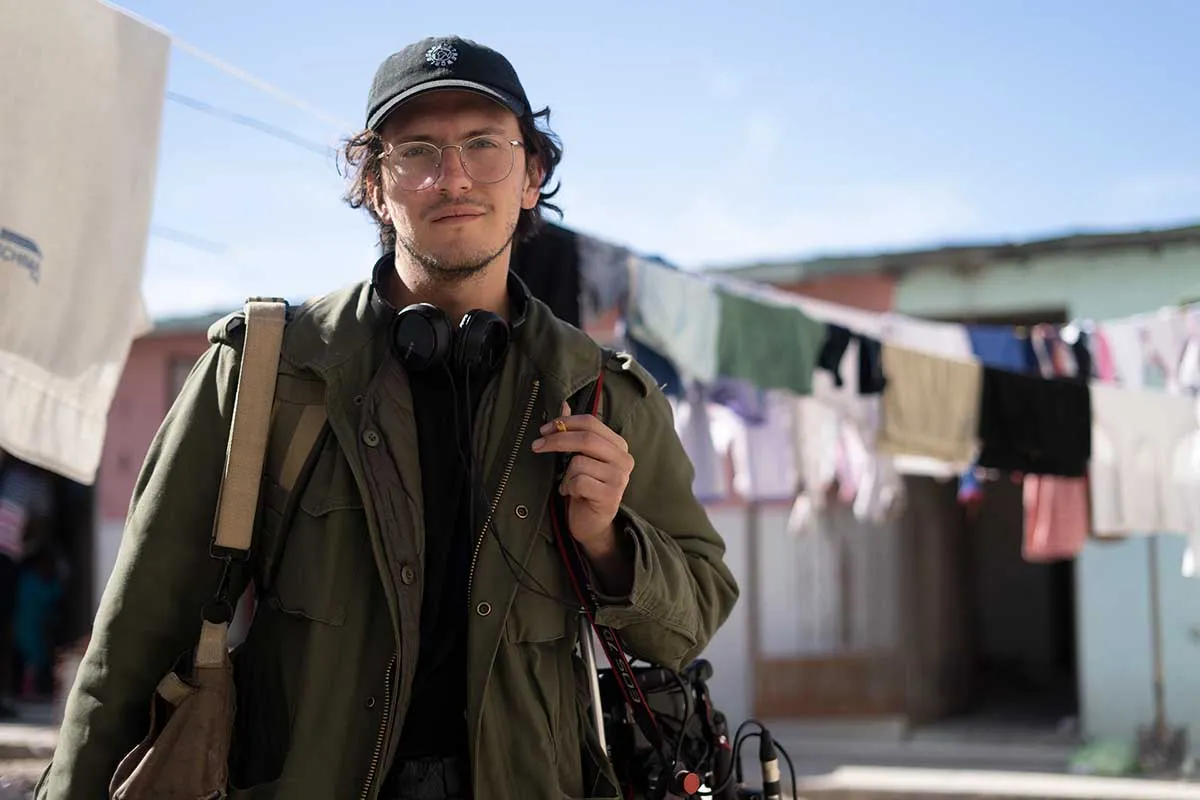 Gerardo del Valle stands outside a migrant house in Acuña, Mexico. He wears a black hat, an olive military jacket and is carrying a video camera and a lens bag. (Photo by Monica Gonzalez)