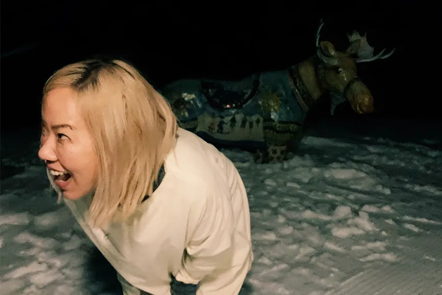 Woman of Korean-descent, she has a big smile and blonde hair to her shoulders. She is looking to her right, in the snow at night with a moose sculpture behind her.