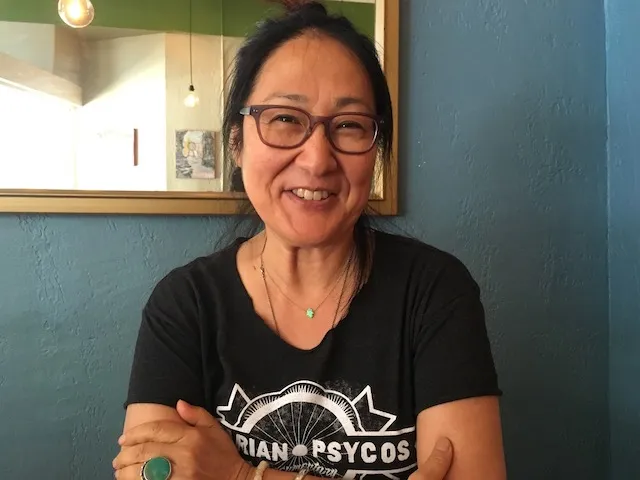 Rea Tajiri, standing in front of a mirror, wearing a black t-shirt, glasses, and a necklace with her hair tied up, smiling.