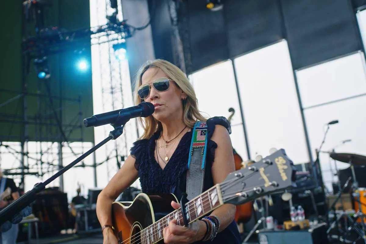 Sheryl Crow, a white female American musician with blonde hair, plays guitar and sings on a large stage. 