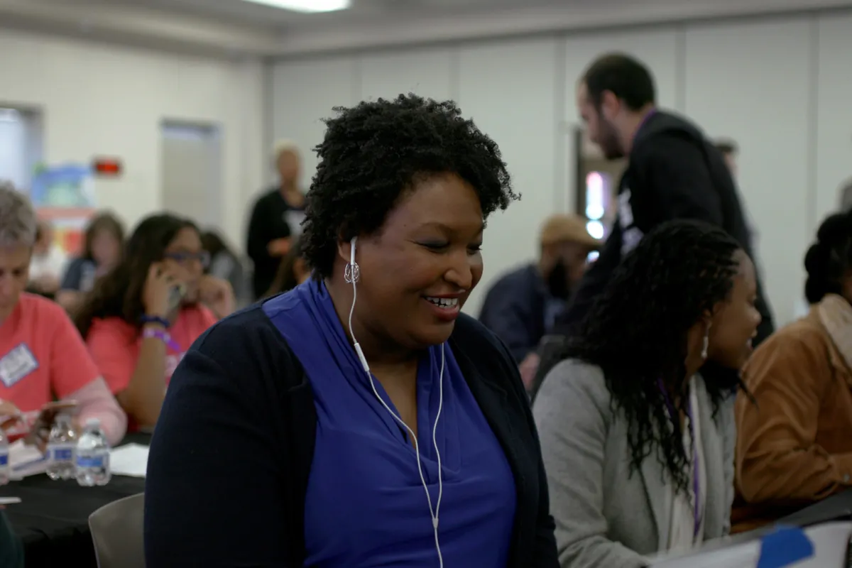 A Black woman in blue shirt and black cardigan, listening to her headphones sitting down
