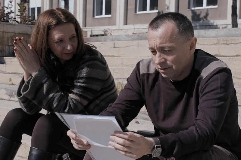 A woman and a man are sitting on the steps of a building, discussing a document