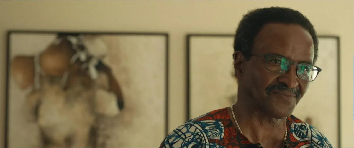 A still image from Descendant, a Black man wearing glasses and a colorful shirt standing in front of a wall inside a house.