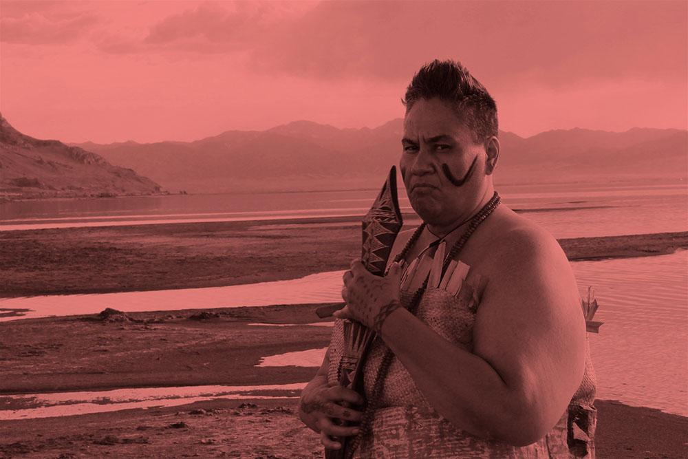 A middle-aged woman with very short hair dressed in traditional Samoan warrior clothes and holding a club near her face and standing at the edge of a lake with mountains in the distance.