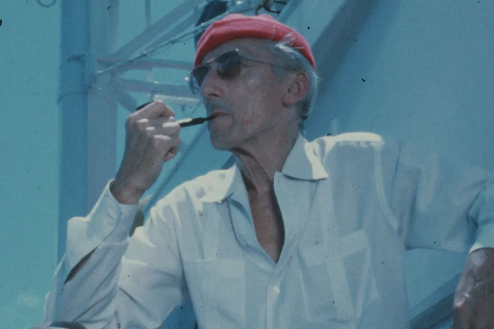 An older white man wearing a red cap, white shirt and sunglasses. He is smoking a pipe.