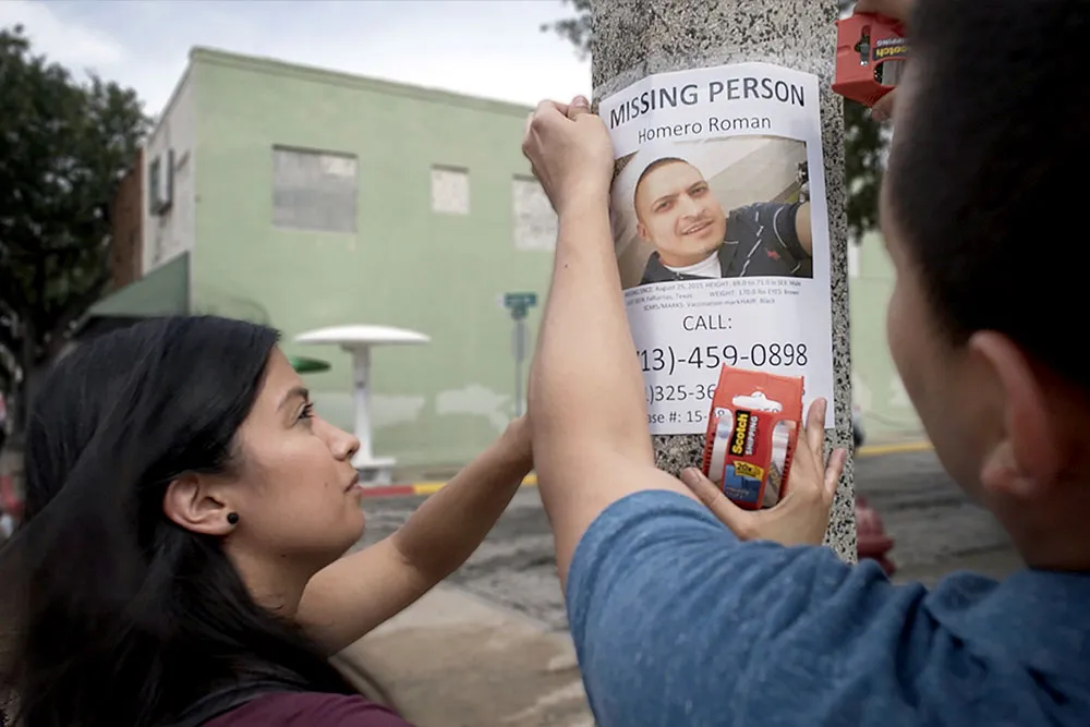 A Latinx woman with black hair and someone off screen hang a poster that reads "MISSING PERSON." It has a picture of a Latinx man with short black hair on it.