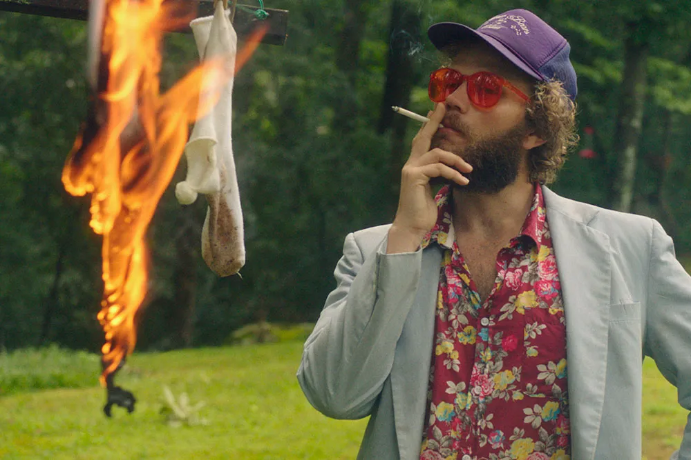 A white man with brown hair and facial hair wearing red shades and a Hawaiian shirt and blazer. He smokes a cigarette and watches socks burn.