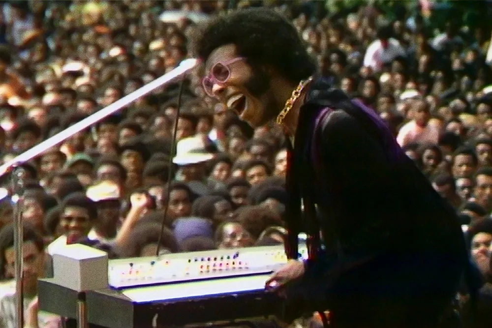 A Black man with black hair wearing pink glasses, a gold necklace and a purple suit. He is playing keyboard to a crowd.