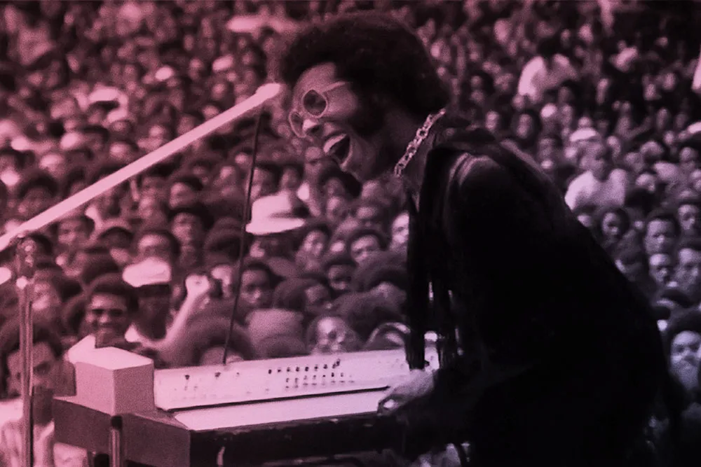 A Black man with black hair wearing pink glasses, a gold necklace and a purple suit. He is playing keyboard to a crowd.