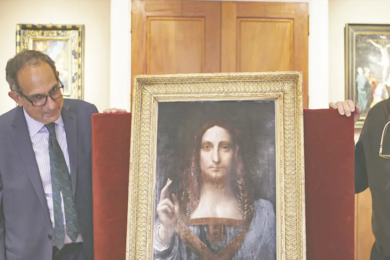 An older white man with black glasses and gray short hair stands next to Salvator Mundi, a painting of a white man with long brown hair pointing upwards.