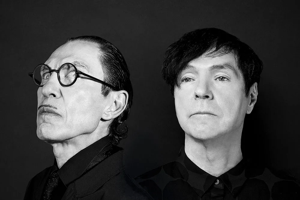 A black and white portrait of two white men both with dark hair. The man on the left has round black glasses and a pencil mustache.