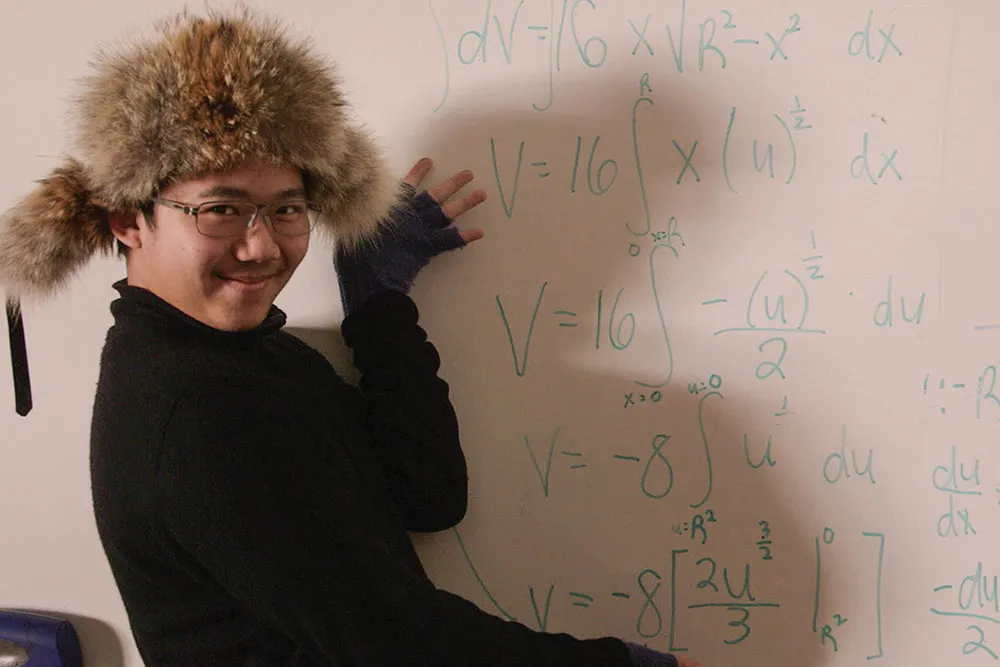 A young Asian student with glasses, wearing a goofy fur hat, motions to a white board with a math formula on it.