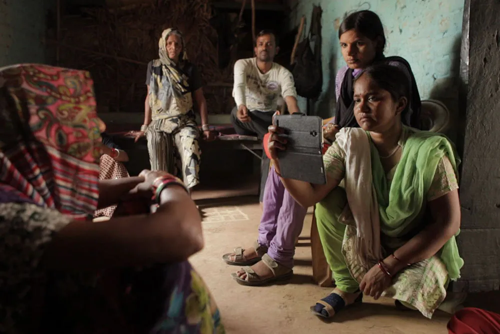 A group of people with brown skin sit in a room. Two women in the front film someone off screen with their phone.