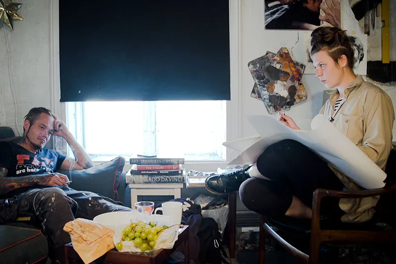 An artist is drawing, her subject sitting across from her on the couch