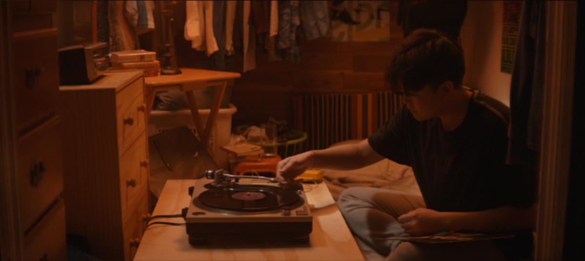 A young guy plays a record player in a small, cramped bedroom.