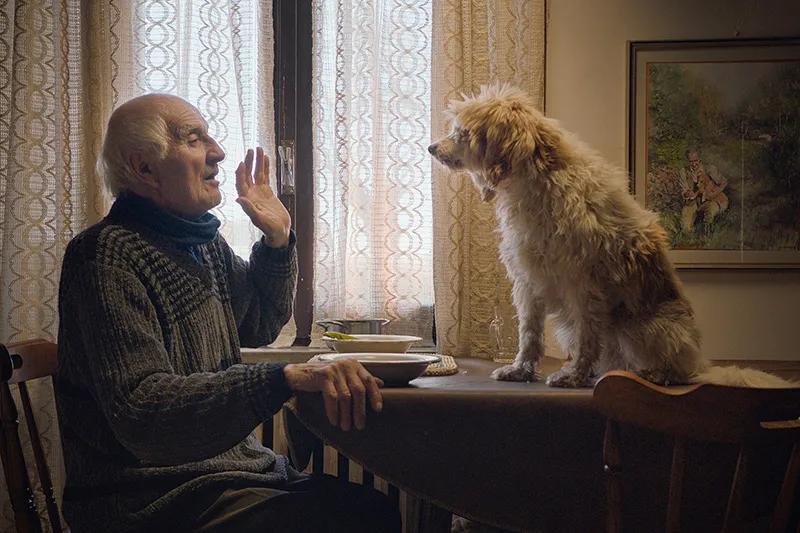 An elder man is talking to his dog that is on the dining table