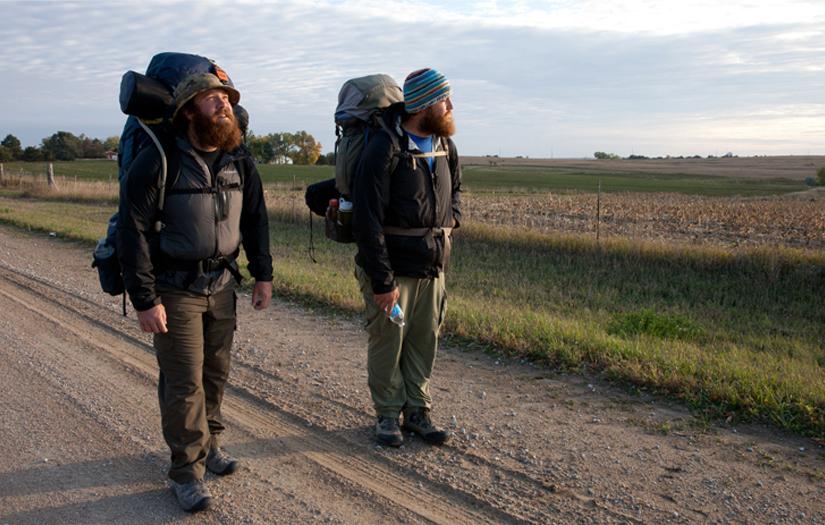 two veterans pause in the middle of road amid their backpacking trip