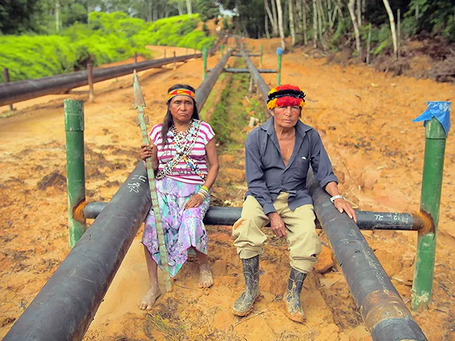 Two indigenous people sitting on black pipes in the Amazonian forest.