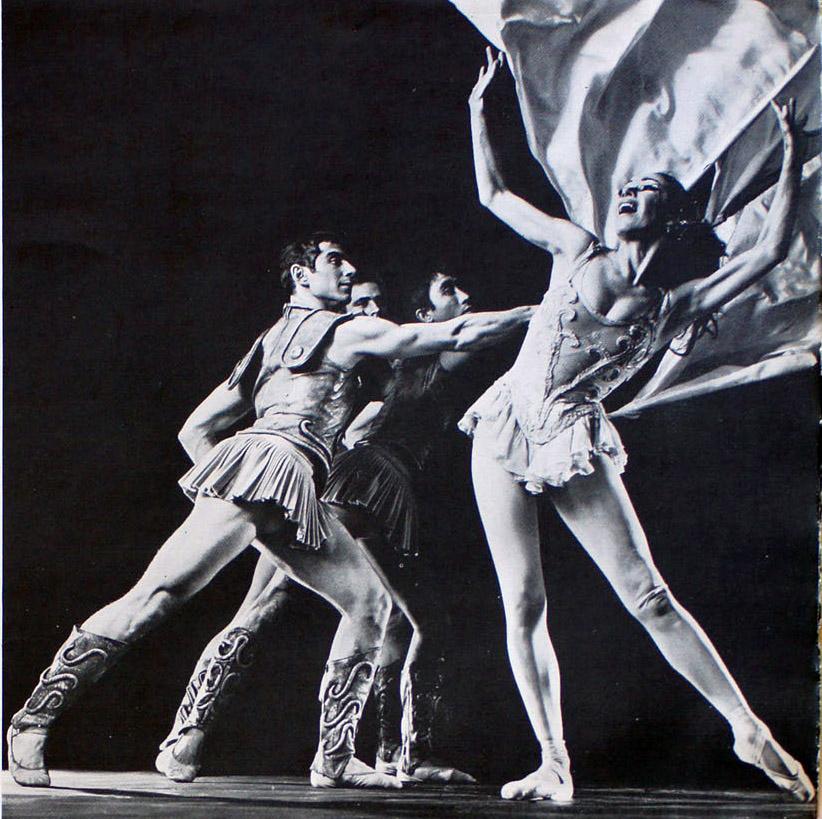 Three White male ballet dancers in Roman Empire guard outfits dance behind a White female ballerina