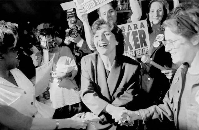 Barbara Boxer is shaking hands with others while supporters hold signs in the background.